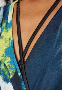 Barevný wrap top s modro-zeleným abstraktním vzorem, s hlubokým výstřihem do V a černými křížovými ramínky zdobenými malými dekoračními cvočky.