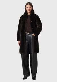 Casaco de pelo sintético preto com gola alta, fecho de botões e bolsos laterais, usado por cima de um suéter escuro e calças de couro. Textura lisa.