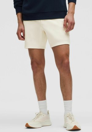 Pantaloni scurți de fleece de culoare crem, cu textură netedă, având un croi clasic și talie elastică, asortați cu șosete albe și adidași deschiși la culoare.