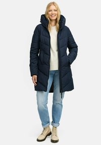 Navy puffer coat met capuchon, voorzien van een ritsluiting en horizontale stiksels. Gepaard met lichtblauwe jeans en beige veterschoenen.