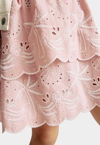 Jupe rose brodée avec des bords festonnés, motifs complexes de bananes, coquillages et fleurs en fil blanc sur un tissu texturé.