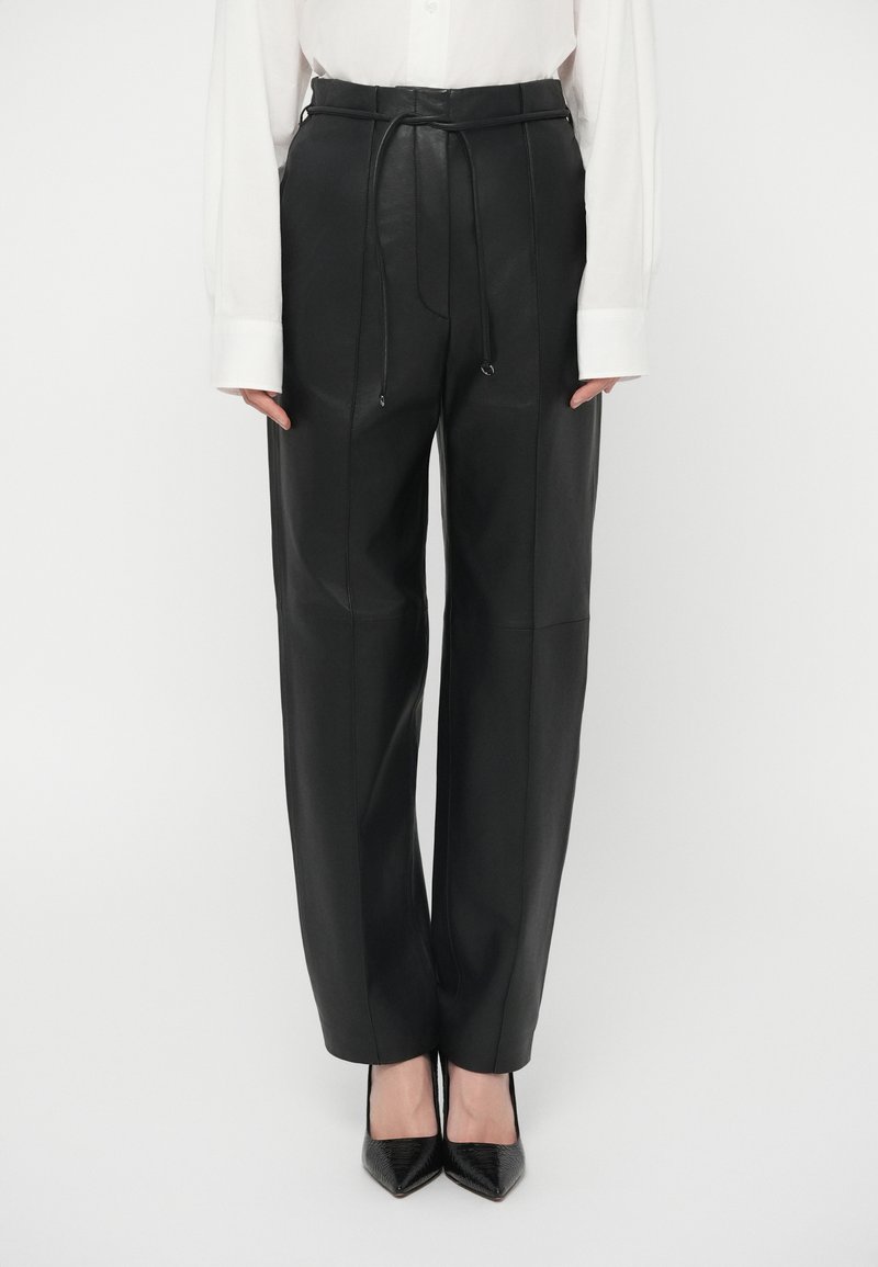 Γυναίκα που φοράει ψηλόμεσα μαύρα δερμάτινα παντελόνια με ζώνη, συνδυασμένα με μαύρες μυτερές γόβες και λευκό πουκάμισο με μακριά μανίκια, που έχει τσιμπηθεί.