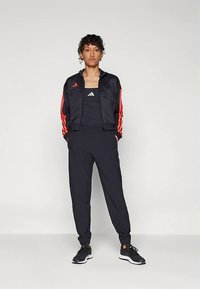 Črna športna jakna s rdečimi trakovi in logotipom, kombinirana s črnimi topom brez rokavov in trenirko, zaključena s črnimi športnimi copati.