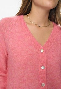 Rozā adīta kardigāna ar V veida kaklu, piecām pogām un mīkstu tekstūru. Iezīmējas ar smalkām krāsu variācijām un vieglu dizainu.