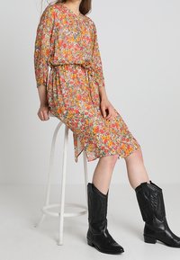 Blommig midiklänning i orange och gula nyanser, tillverkad av lätt tyg med ett bälte i midjan, i kombination med svarta cowboystövlar.