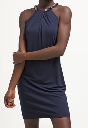 Vrouw draagt een mouwloze marineblauwe jurk tot op de knie met een met kralen versierde halternek, staand met één arm over de andere gekruist.