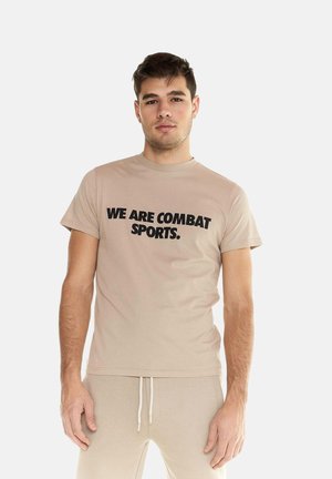 Jonge man met beige T-shirt met de tekst "WE ARE COMBAT SPORTS." en bijpassende beige broek, staand tegen een effen lichte achtergrond.