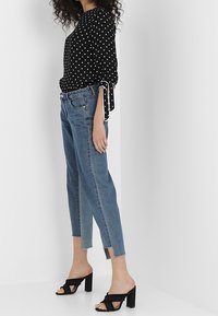 Černá halenka s bílými puntíky, dlouhé rukávy, volný střih. Světle modré džíny s rovnými nohavicemi a neupraveným lemem. Černé sandály na podpatku se širokými pásky.