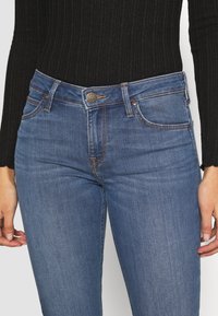 Jeans de mezclilla azul con un diseño ajustado, un desvanecimiento sutil y costuras en contraste, con un cierre de botón etiquetado como "Lee" en la cintura.