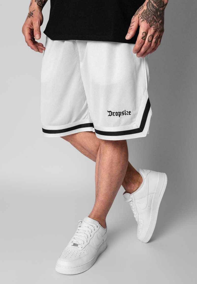 Homme portant un short de basketball blanc Dropsize avec un liseré noir et des baskets blanches, debout les jambes croisées sur fond gris.