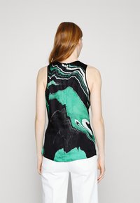 Mujer con cabello rojo, vestida con una blusa negra sin mangas con un patrón abstracto en verde y blanco, combinada con pantalones blancos, de pie frente a un fondo liso.