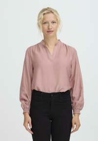 Rosa Langarmbluse mit V-Ausschnitt, strukturiertem Stoff und gerafften Manschetten, kombiniert mit schwarzen engen Hosen.