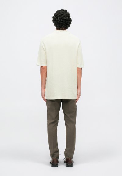 Svetlo bež srajca s kratkimi rokavi in teksturiranim vzorcem, v kombinaciji s sivimi hlačami in rjavimi čevlji, prikazana s hrbtne strani.