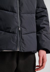 Mörk pufferjacka med en slät, texturerad yta, utrustad med en central dragkedja och quiltade partier för isolering. Hand placerad på sidan.