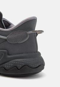 Šedá športová obuv s textúrovanou gumovou podrážkou, hladkým textilným vrchom a charakteristickou zadnou nášivkou. Zaoblený tvar so šitými akcentmi.