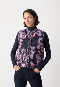 Gep quiltete schwarze Weste mit pinken Blumenmustern, rundem Kragen, Vorderknöpfen und zwei großen Taschen, getragen über einem marineblauen Langarmoberteil.