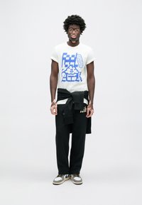 Balti marškinėliai su mėlynu grafiniu dizainu, juodos Nike logotipo laisvalaikio kelnės ir pilki/balti sportbačiai. Modelis turi trumpus garbanotus plaukus.