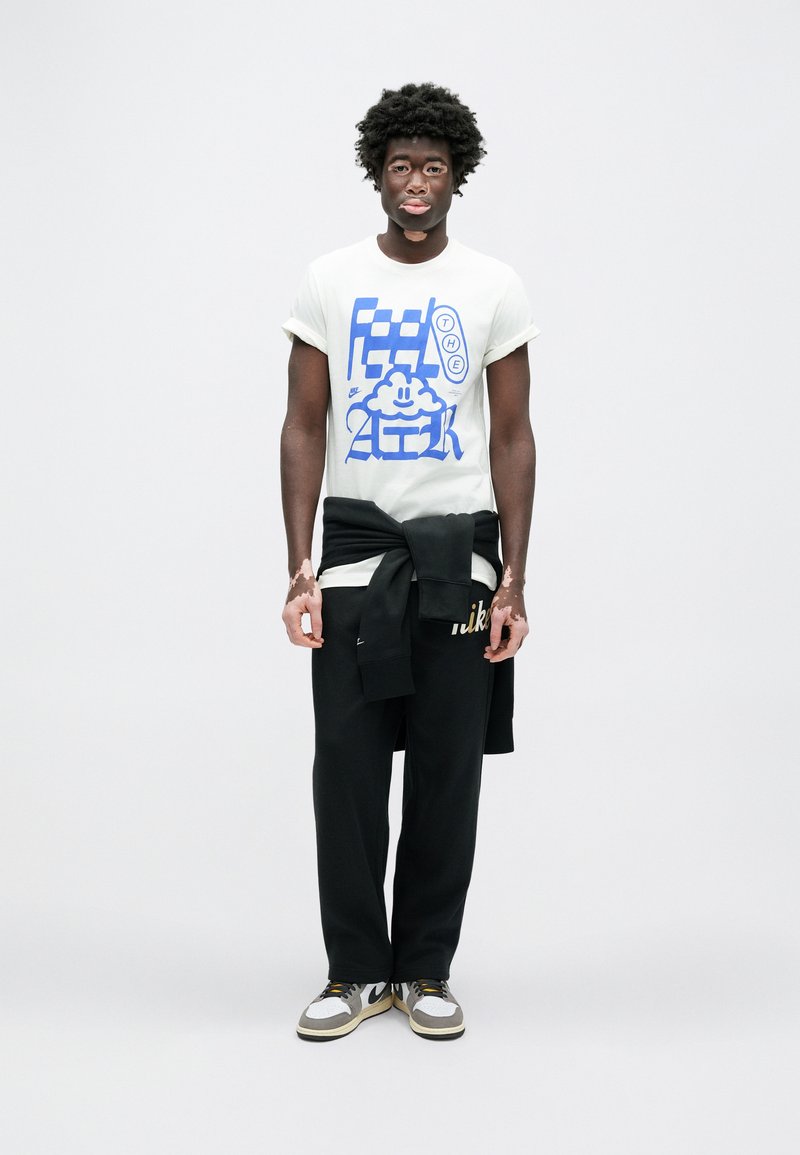 T-shirt blanc avec un motif graphique bleu, pantalon de survêtement noir avec logo Nike, et baskets grises/blanches. Le modèle a des cheveux courts et bouclés.