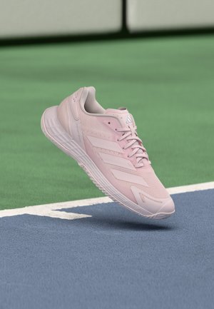 Lichtroze Adidas sportschoen zweeft boven een blauwgroen tennisveld met witte begrenzingslijnen.