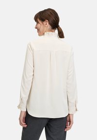 Crème blouse met een gerimpelde kraag en manchetten, met een achterste plooi en knoopmanchetten, gemaakt van een gladde stof. Achteraanzicht weergegeven.