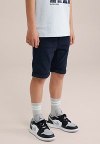 Tamsiai mėlyni džinsiniai šortai su pasuktais kraštais, derinami su šviesiai pilkomis kojinėmis, turinčiomis dvi baltas juosteles, ir juodais bei pilkais sportbačiais.