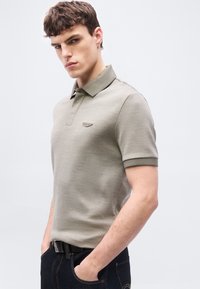 Νεαρός άνδρας που φοράει μπεζ κοντομάνικο πόλο και σκούρο τζιν με τα χέρια στις τσέπες, στέκεται μπροστά σε μονόχρωμο φωτεινό φόντο.