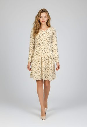 Mujer joven de pie, vistiendo un vestido beige de manga larga con pequeñas manchas multicolores y tacones altos crema sobre un fondo claro y sencillo.