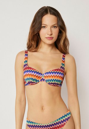 Kvinde med langt, bølget hår iført farverig bikini med zigzag-mønster på top og bund mod en ensartet lys baggrund.