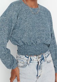 Niebieski krótki sweter z dzianiny z długimi, bufkami rękawami i elastycznym, marszczonym pasem, noszony z jasnymi, wysokim stanem szortami jeansowymi. Widoczny łańcuszkowy naszyjnik.