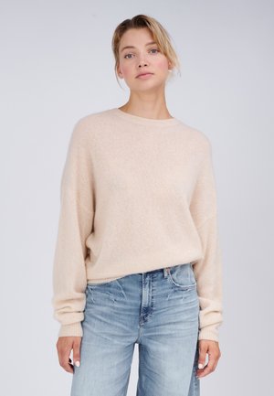 Frau mit hellem Haar, die einen beigen Strickpullover und hellblaue Jeans mit hoher Taille trägt, steht vor einem schlichten hellen Hintergrund.