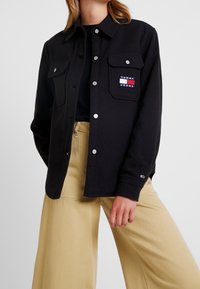 Czarna kurtka koszulowa z fakturowanej tkaniny, z srebrnymi napami i kieszenią na piersi z naszywką logo "Tommy Jeans". Połączona z beżowymi spodniami w szerokim kroju.