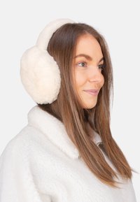 Cache-oreilles en fausse fourrure blanche avec un design arrondi, une texture douce et un bandeau ajustable, portés sur une veste à col en peluche blanche.
