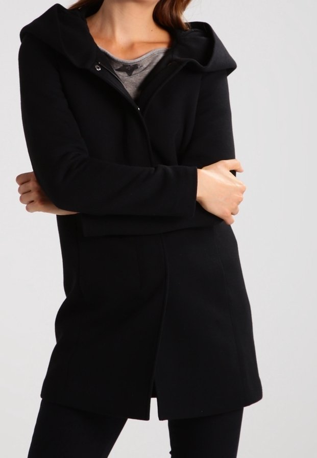 Abrigo negro con capucha y cierre de cremallera, hecho de una tela suave. Presenta una silueta ajustada y bolsillos laterales. Llevado sobre una blusa gris.