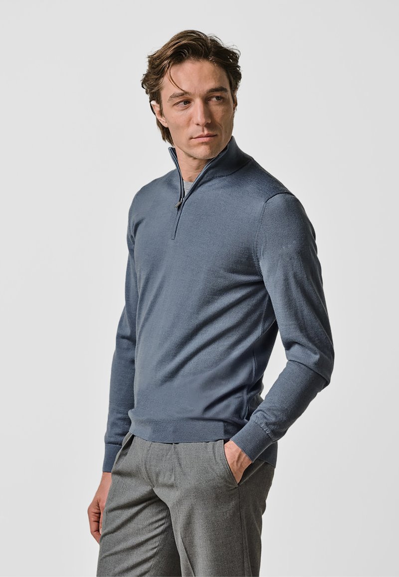 Pull-over zippé pour homme en bleu chiné, fabriqué en tissu tricoté. Il présente un col montant et une coupe ajustée, associé à un pantalon gris clair.