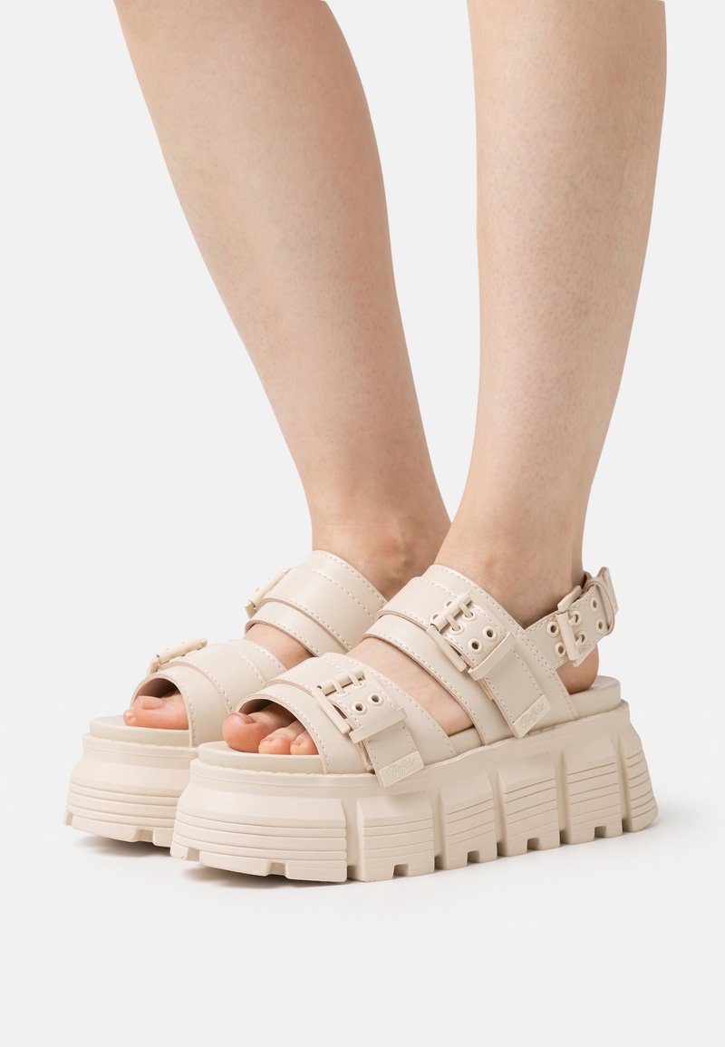 Pieds portant des sandales compensées beiges avec des semelles épaisses rainurées, des brides réglables et des boucles sur un fond clair uni.