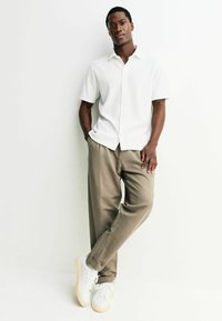 Weißes Kurzarm-Hemd mit durchgehender Knopfleiste und geripptem Stoff, kombiniert mit locker sitzenden beigen Hosen und weißen Sneakers mit hellbeigen Sohlen.