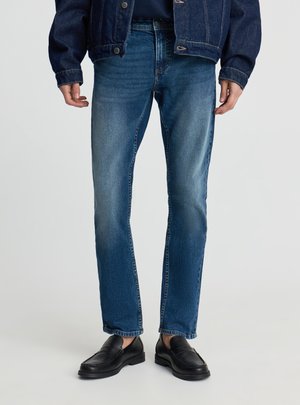 Uomo che indossa jeans a gamba dritta di media tonalità, mocassini neri in pelle e una giacca di denim scuro su uno sfondo chiaro e semplice.