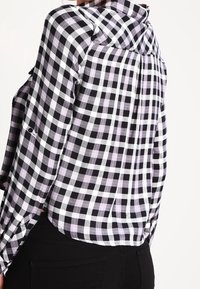Persona che indossa una camicia a maniche lunghe a scacchi neri, bianchi e lavanda abbinata a pantaloni neri, vista da dietro e di lato.