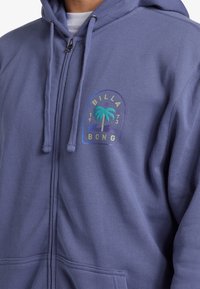 Felpa con zip in tessuto viola chiaro con una grafica di palma e il testo "BILLA" e "BONG". Presenta lacci e tasche frontali.