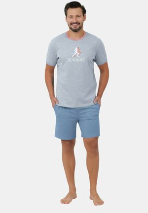 Uśmiechnięty mężczyzna stoi boso, ubrany w szary t-shirt z logo "RUNNING" oraz jasnoniebieskie szorty, ręce w kieszeniach.