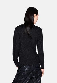 Zwarte trui met een gemêleerd breisel en subtiele glans. Heeft lange mouwen en een ronde halslijn. Ggedragen met glanzende zwarte broeken.
