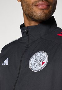 Veste de sport noire avec un col haut, présentant des accents blancs et rouges, logo brodé sur la poitrine et tissu texturé. Devant zippé.