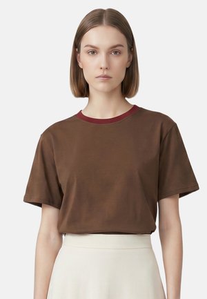 Femme aux cheveux lisses mi-longs, portant un t-shirt marron avec un col rouge et une jupe blanche, regardant droit devant avec une expression neutre.