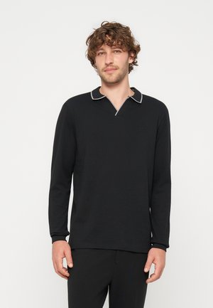 Homme aux cheveux bouclés portant une chemise noire à manches longues avec un col bordé de blanc, debout devant un fond clair uni.