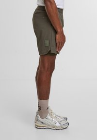 Bermudas de color verde oliva con un acabado texturizado, que cuentan con un bolsillo lateral y una etiqueta rectangular. Combinadas con zapatillas deportivas plateadas y calcetines beige.