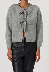 Cardigan grigio lavorato a maglia con due legature frontali, scollo rotondo e polsini a coste. Indossato con pantaloni neri aderenti, che mettono in risalto un design corto.