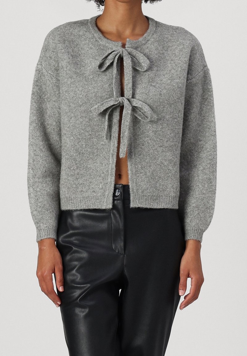 Cardigan grigio lavorato a maglia con due legature frontali, scollo rotondo e polsini a coste. Indossato con pantaloni neri aderenti, che mettono in risalto un design corto.