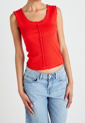 Femme portant un haut rouge sans manches avec des bords volantés et un jean taille haute bleu clair sur un fond uni.