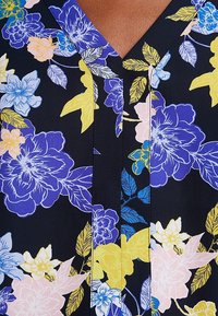 Kvetinová blůza s výstřihem do V, zdobená barevnými květy v modré, růžové a žluté barvě na tmavě navy pozadí, předvádějící hladkou texturu látky.