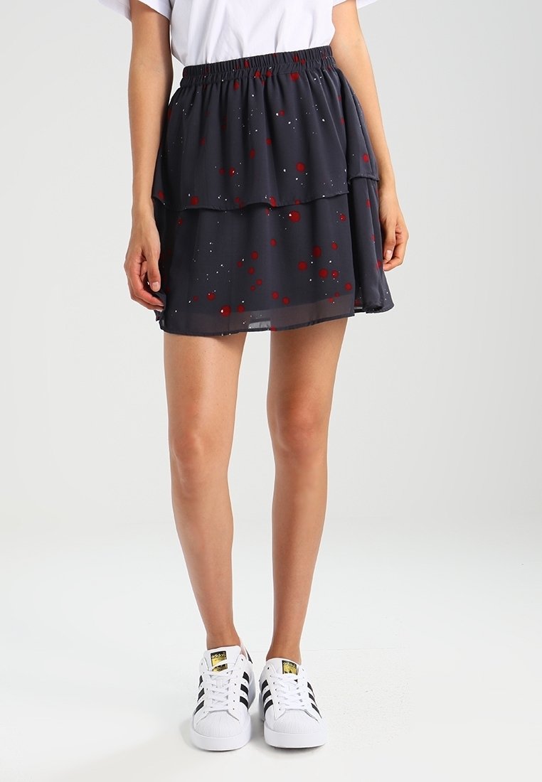 Femme portant une jupe noire superposée avec des motifs éclaboussés rouges et blancs, des baskets blanches avec des rayures noires et un haut blanc, debout.
